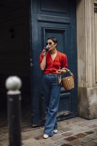 A woman at Paris Fashion Week wearing a red cardigan sweater, blue jeans, flats, and a tan basket bag