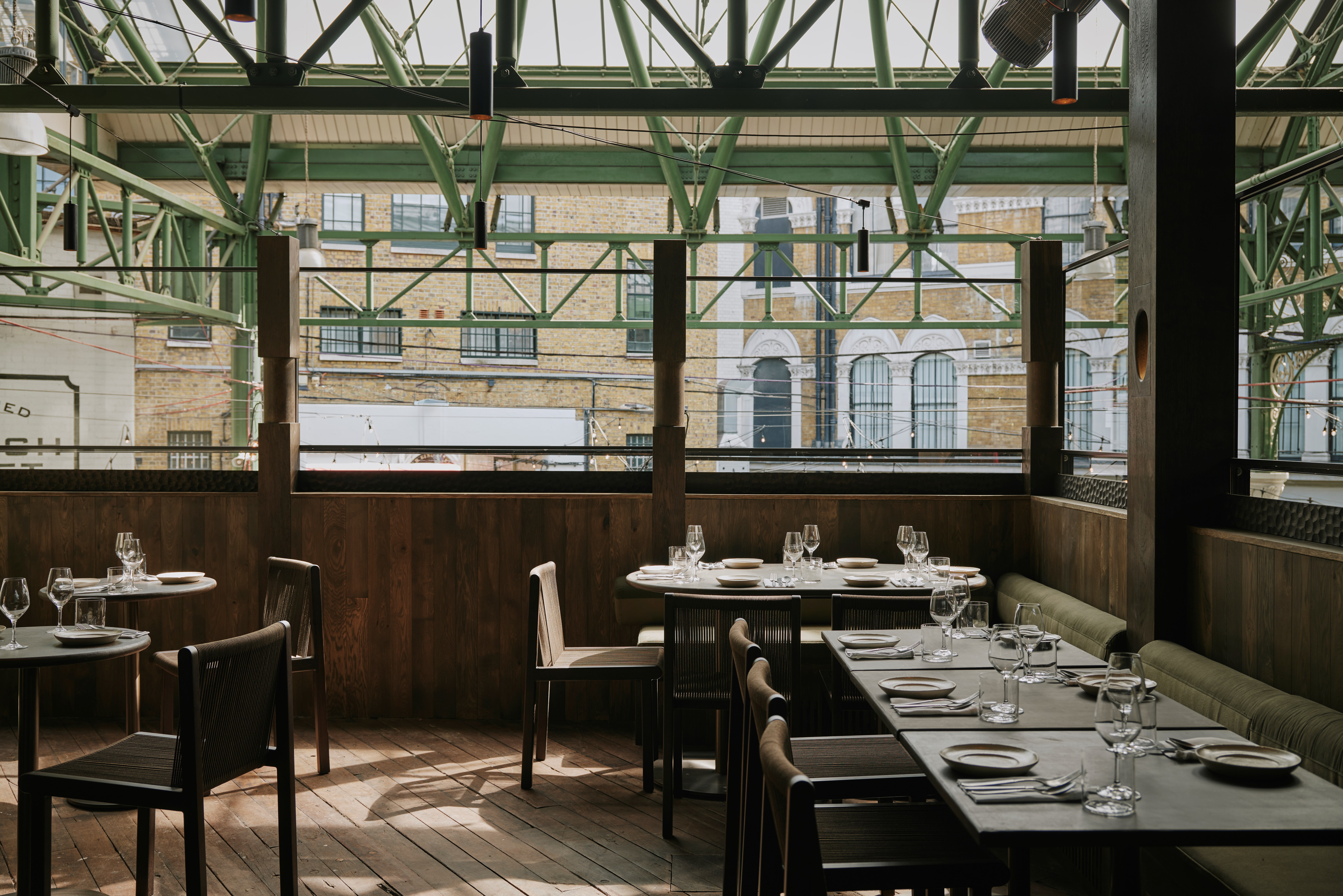 A terrace of outdoor seating with wooden chairs on a wooden flooring with wooden balcony. Overhead, pale green Victorian ironwork forms triangles of scaffolding.
