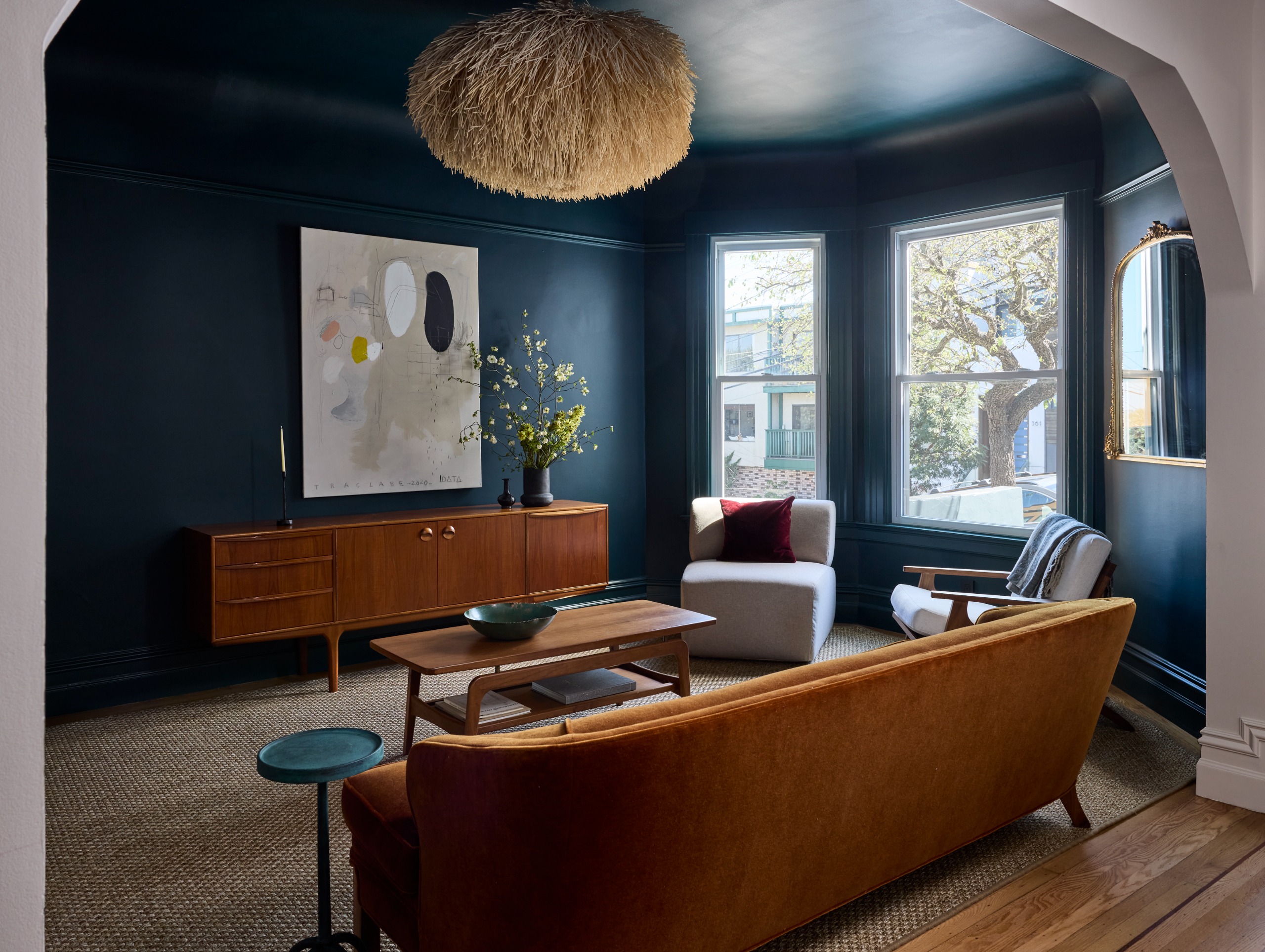 A dark blue living traditional room with a mid century sideboard and coffee table, armchairs and sofa around the coffee table with artwork as the focal point.