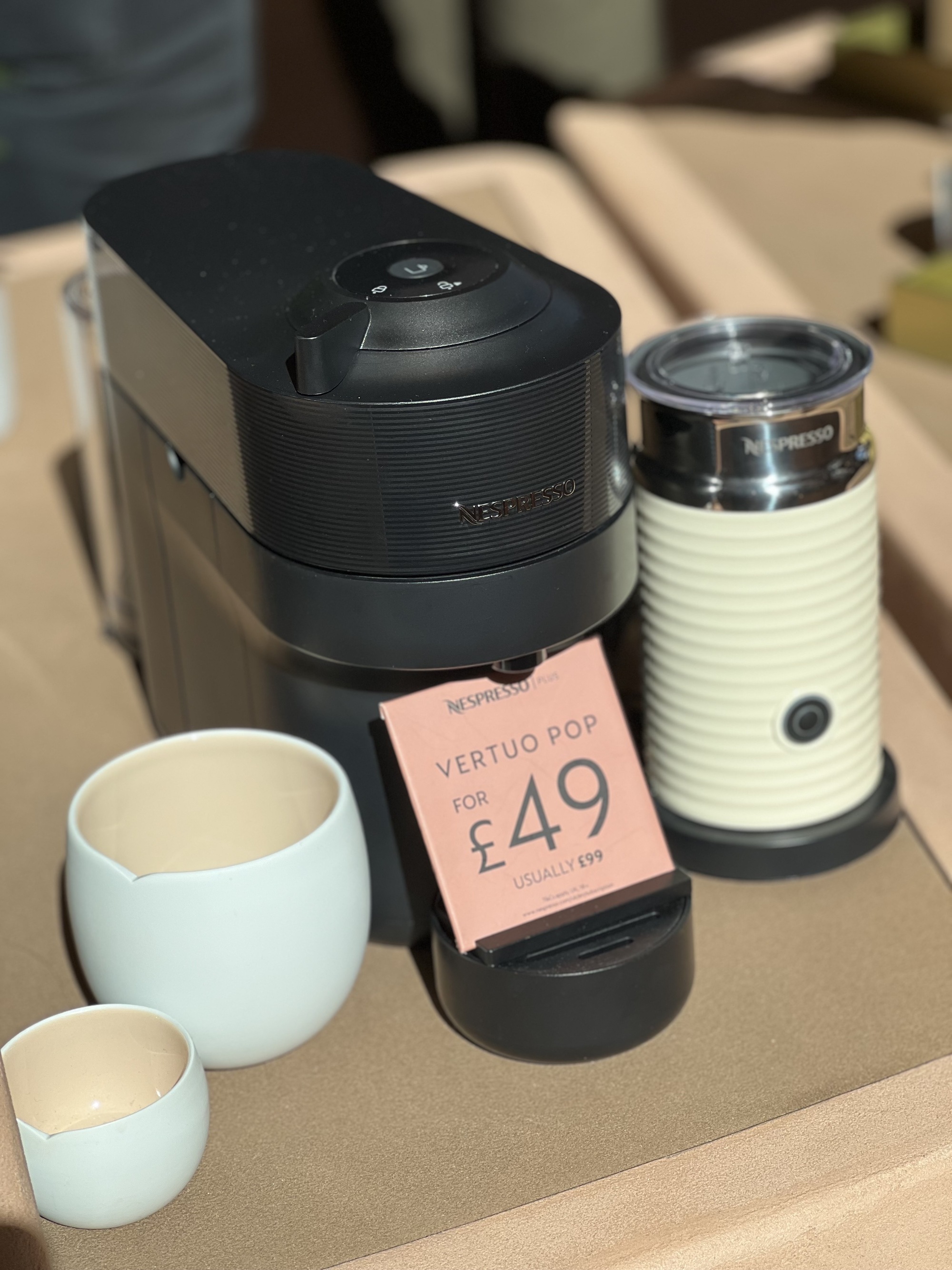A black coffee pod machine with a white milk frother and two white ceramic mugs next to it in a sunny showroom.