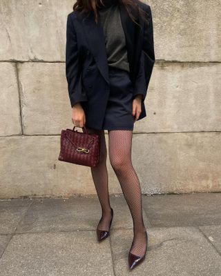 Woman carrying a burgundy Bottega Veneta bag and wearing a black skirt suit, fishnets, and pointed-toe heels.