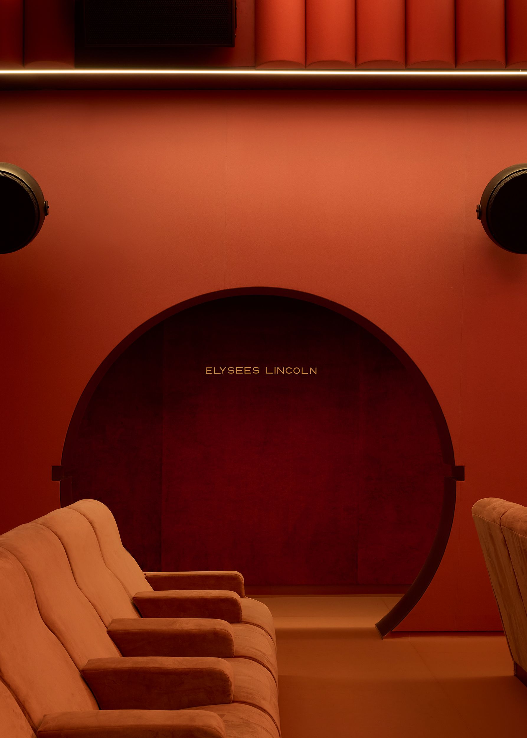 A cinema screening room with velvety red seats, porthole window-like ceiling lights, high-tech sound panels in pink, rounded shapes, and semi-circular portals.