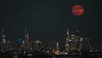 A red full moon rises in a dark sky above a city skyline crowned by skyscrapers.