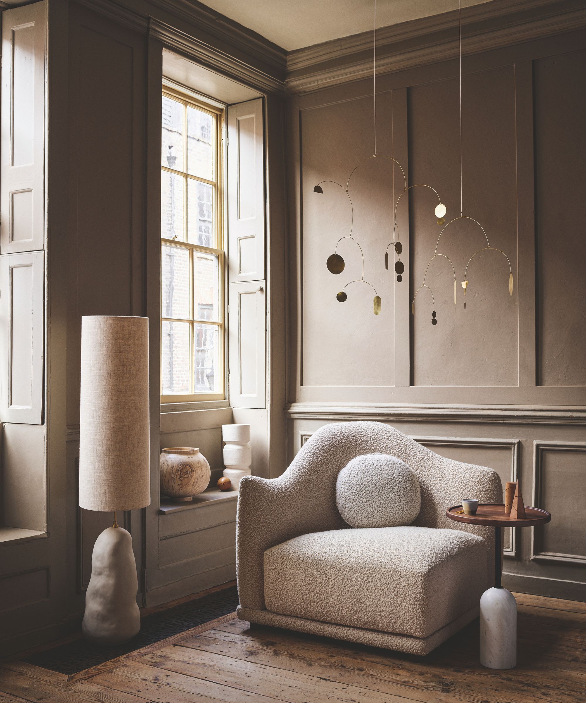 A brown panelled living room with hardwood floors, a boucle chair, large windows with shutters, a white lamp and circular table. Hanging from the ceiling is a gold mobile.