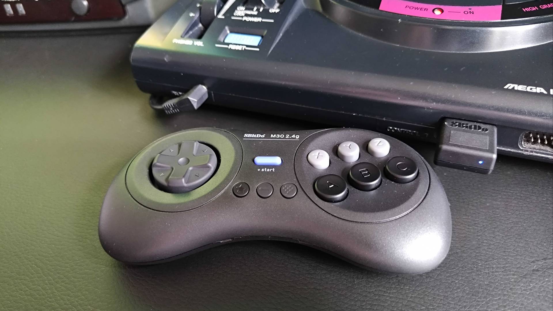 8Bitdo M30 2.4Ghz wireless Sega Mega Drive/Genesis controller next to Japanese console on black desk with adapter inserted into controller port.