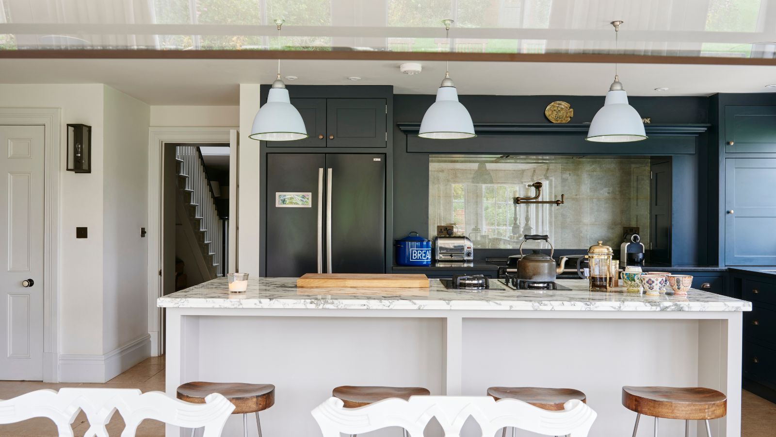 A white and black kitchen with black cabinets, a white island with marble countertops and white pendant lights hanging above, wooden barstools and white chairs in the foreground. 