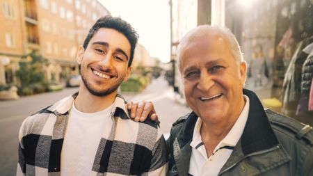 A smiling grandfather and grandson stand on a street. The young man is in his early 20s. The grandfather has his hand on his shoulder and looks protective.