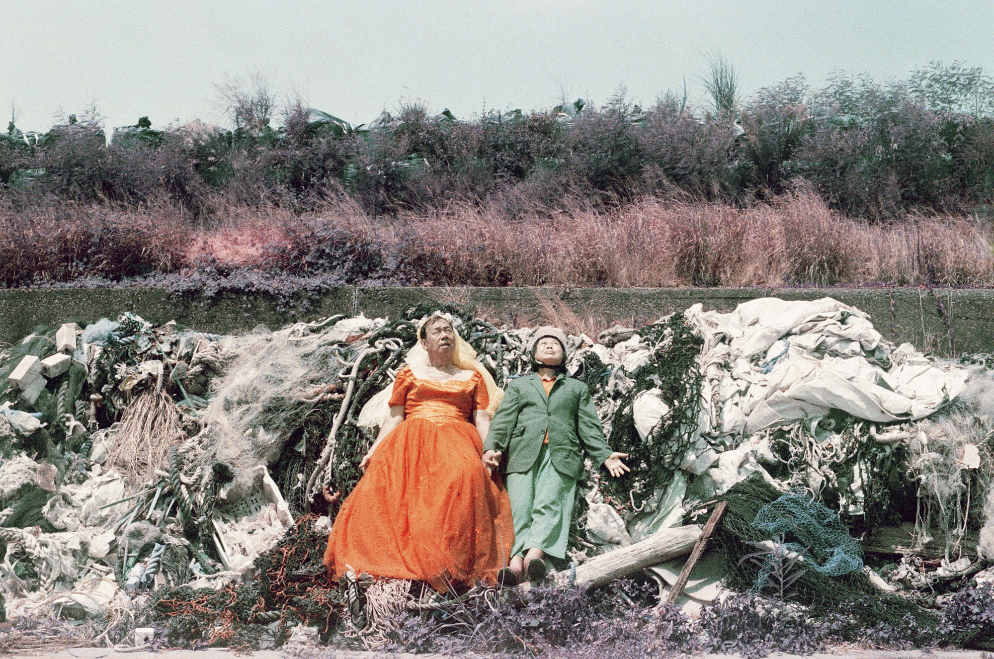 A man in a bright orange dress and a woman in a green suit lie side-by-side with closed eyes on a large pile of plastic waste and discarded fishing nets outdoors.