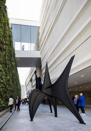 Close up exterior view of The San Francisco Museum of Modern Art (SFMoMA) during the day. There are a number of people outside along with a wall of greenery and a black structure entitled 'Trois Disques' ('Three Disks') by Alexander Calder