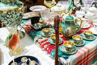 A number of gold and turquoise-decorated ceramic mugs and teapots, silver mugs and other sculptural home accessories sit on a table covered with a chequered throw at a flea market.