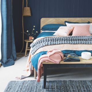 coastal bedroom ideas with navy painted tongue and groove, rope rattan light, pale wood bed, blue and blush bedding, grey knitted throw, pink cushions, weathered bench, blue textured rug, denim curtains
