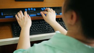 A close-up shot of a vision impaired person using a computer keyboard and braille display, to represent accessibility in tech. The person is shot from behind and is wearing smart casual clothing.