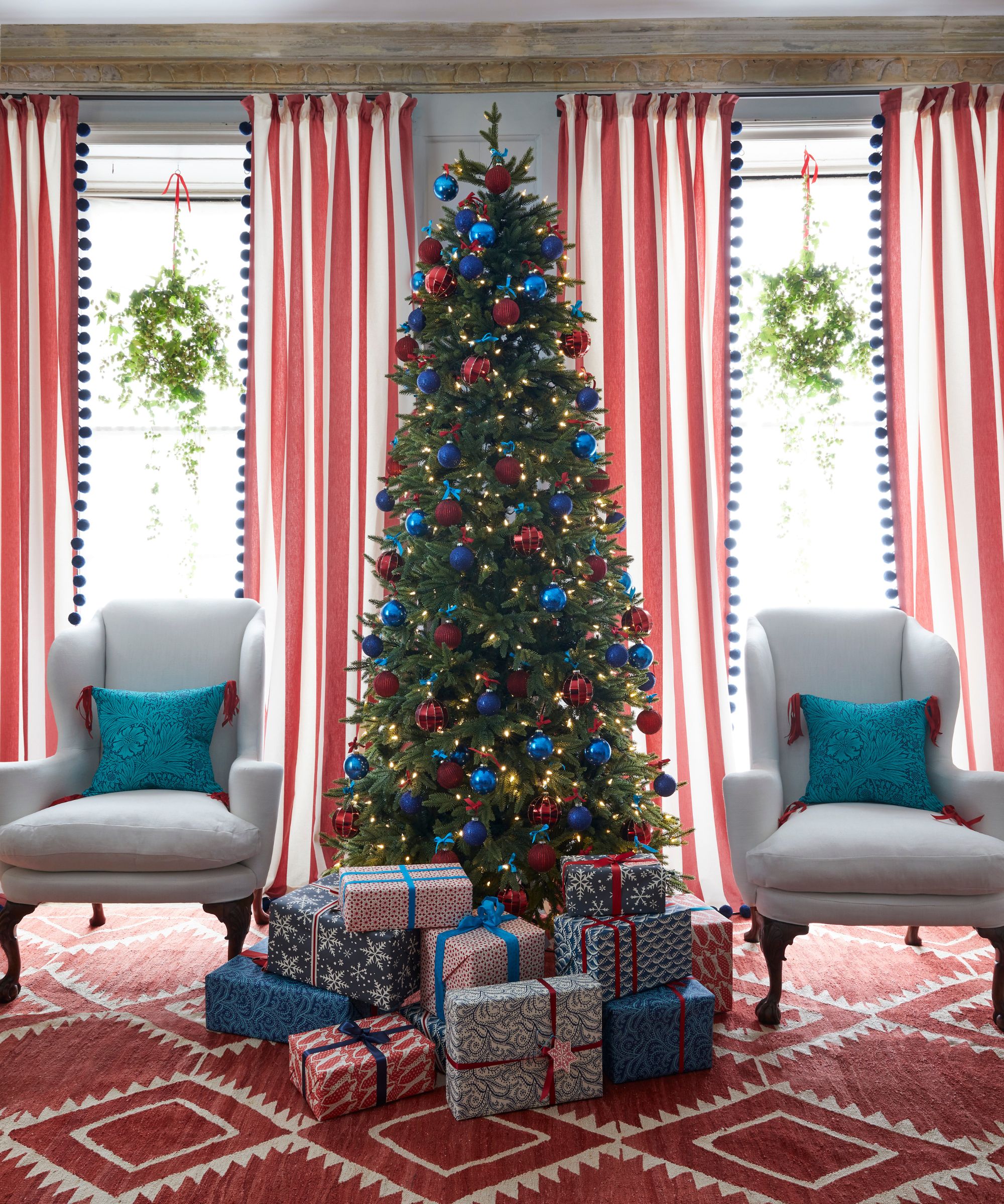 A festive red living room with a tall, narrow decorated Christmas tree with blue and red baubles and yellow lights with wrapped gifts underneath, two white upholstered chairs with blue cushions, and two large windows to either side of it with red and white patterned curtains with wreaths. The carpet is red and white patterned.