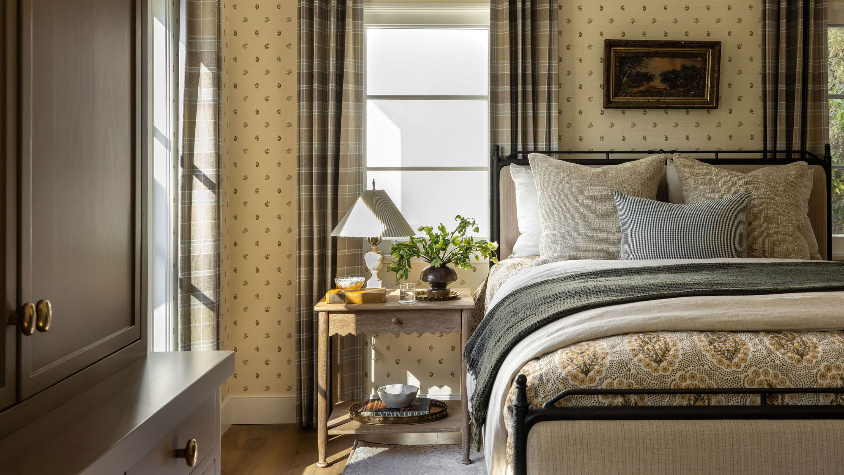 a layered bedroom in an ADU with floral wallpaper, metal bed, vintage art and plaid drapes