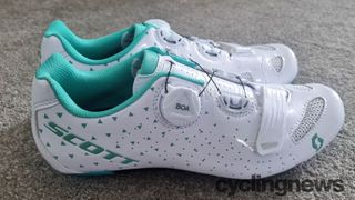 A pair of white and turquoise Scott Road Comp Boa Lady Shoes side by side on a grey carpet