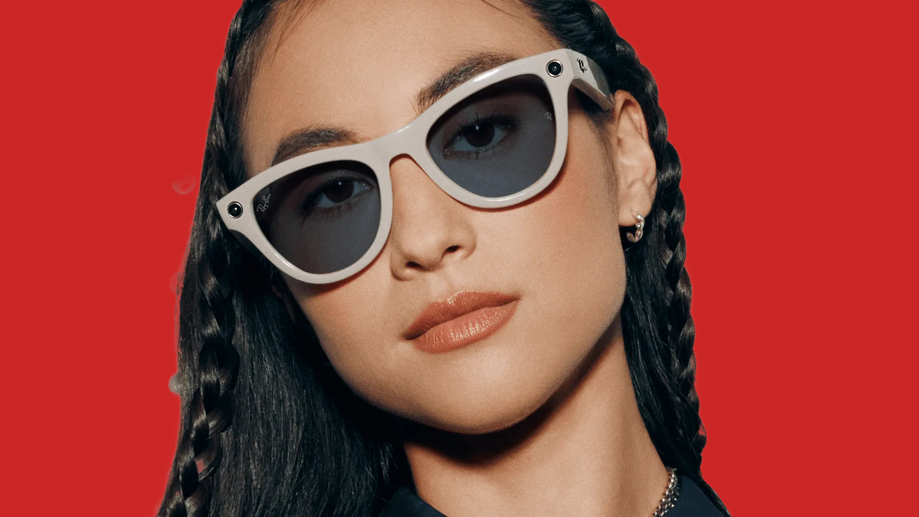 A woman wearing a pair of Meta Ray-Ban smart glasses looking directly at the camera in front of a red background.