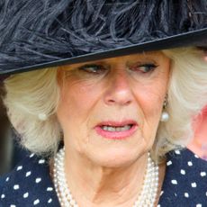 Queen Camilla wears a polka dot dress, a pearl necklace and black hat with feathers in it and she looks tearful