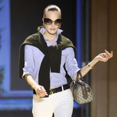 Model walks the Gucci SS27 Runway wearing jodhpur leggings tucked into a shirt with a jumper over the shoulder and black knee-high boots.