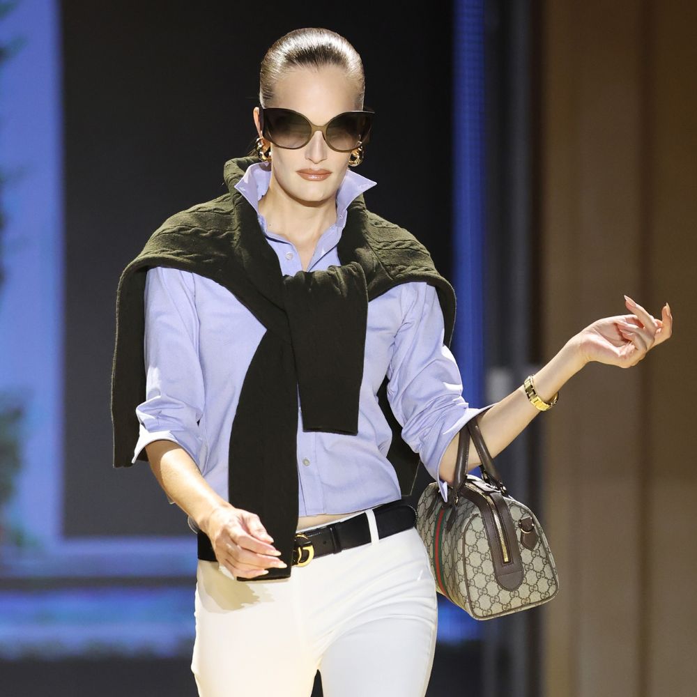 Model walks the Gucci SS27 Runway wearing jodhpur leggings tucked into a shirt with a jumper over the shoulder and black knee-high boots.