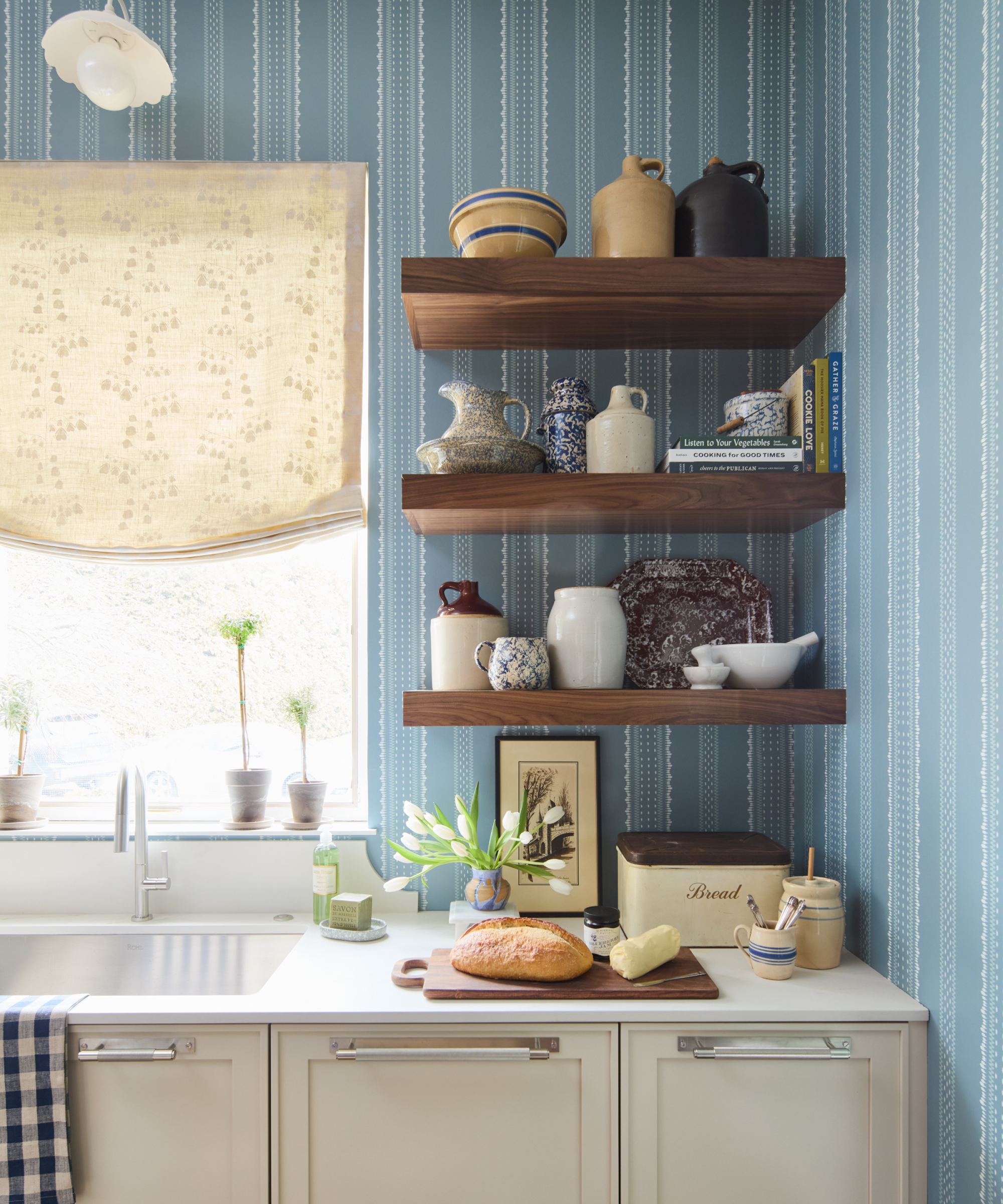 off white kitchen units with blue striped wallpaper on walls, small dark wood shelves and cream blind at window above sink