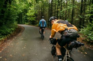 The Jack Wolfskin bikepacking range of clothing on two riders on is looking back at the camera another is in front with hands off the bars on a road with trees either side of the road