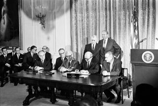 a group of men in suits sign a document at a wooden table in front of the american flag