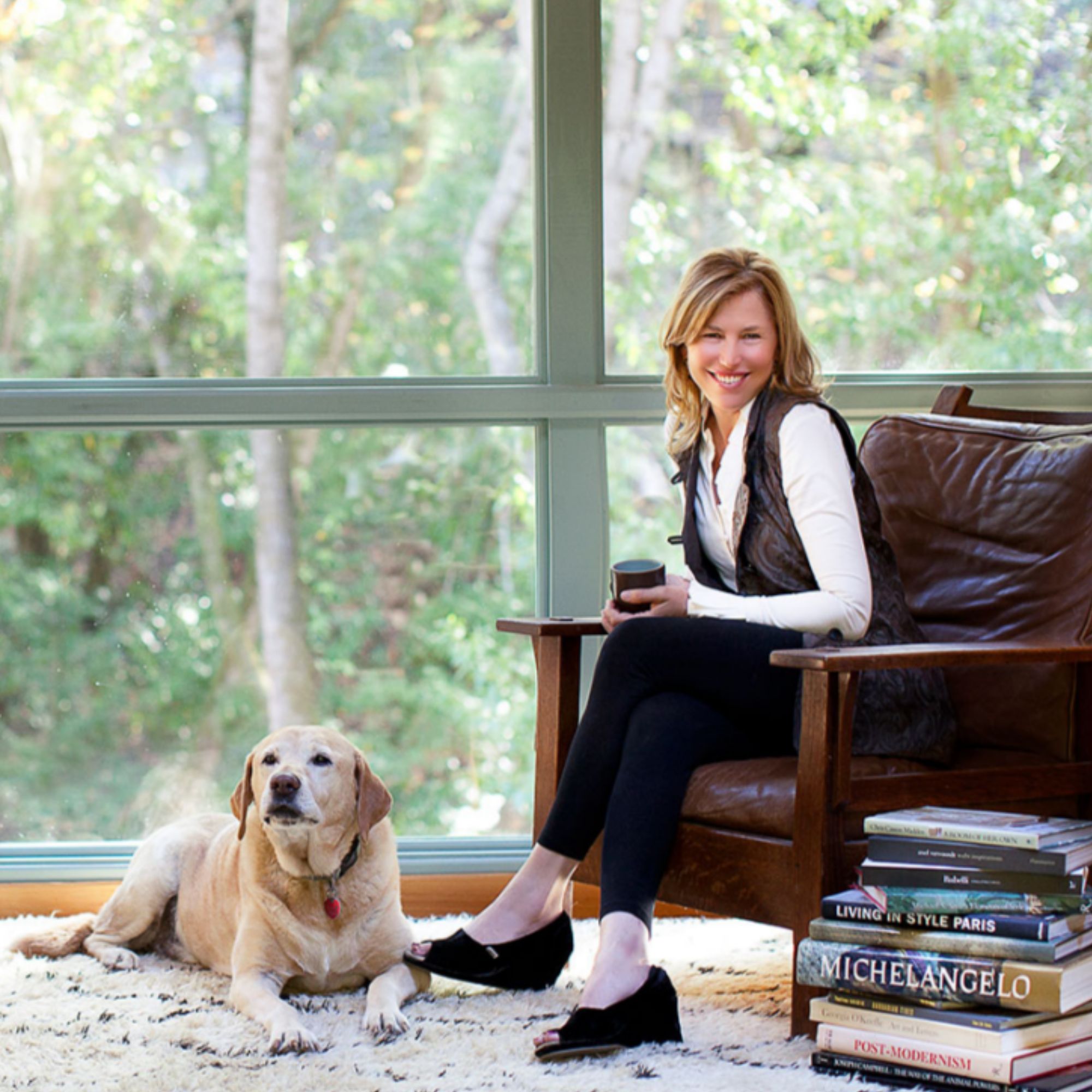 A headshot of designer Lisa Staprans, sitting in a chair with a dog on the floor