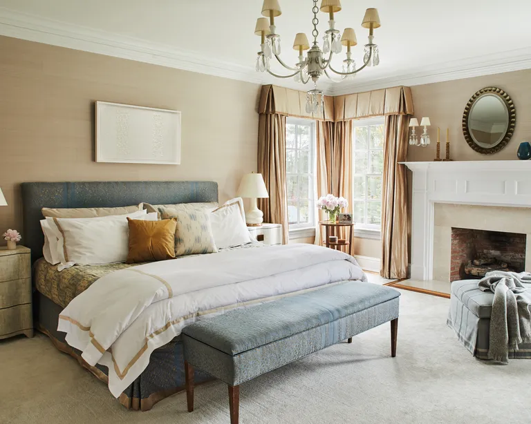 A bedroom with blue upholstered bed and storage bench, sand walls and white fireplace