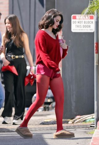 A photo of Selena Gomez sporting a cherry red sweater, shorts, and sheer tights with platform shearling slides.