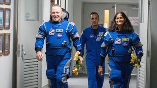 A photo of two astronauts in blue jumpsuits walking down a hallway with another man behind them