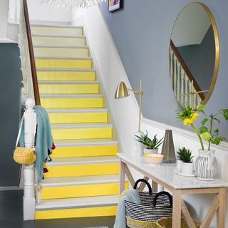 White stair case with yellow steps next to wooden hallstand with granite top with round copper framed mirror above it