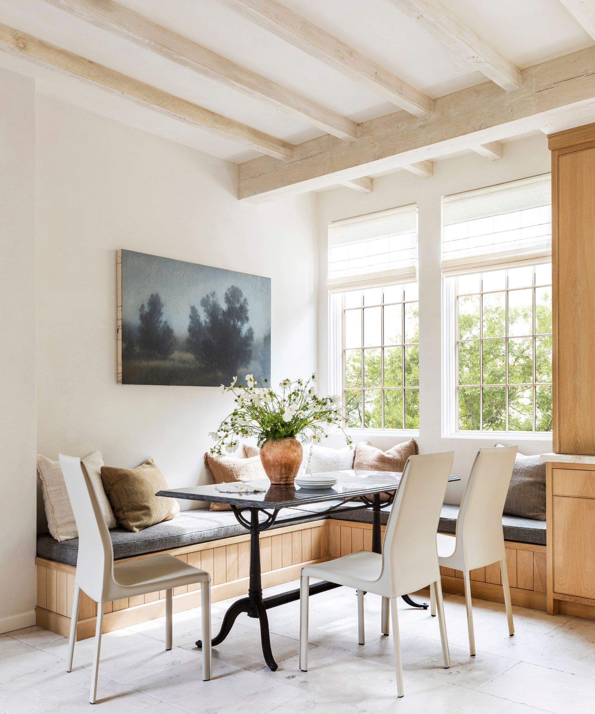 Breakfast nook with white walls, beamed ceiling, window seat and banquette seating, wooden cabinetry, a black table and white dining chairs with a forest landscape picture on the wall above.