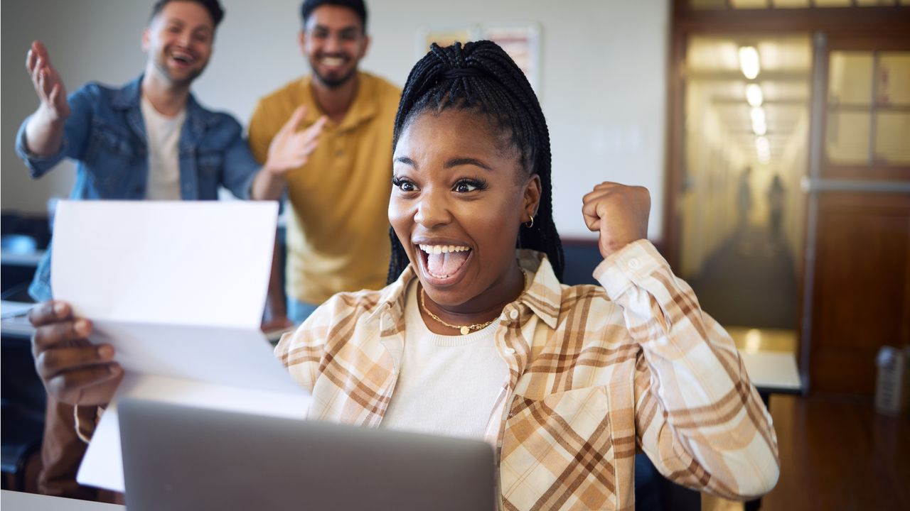 A woman sitting at her desk celebrates some good news while co-workers look on in the background.