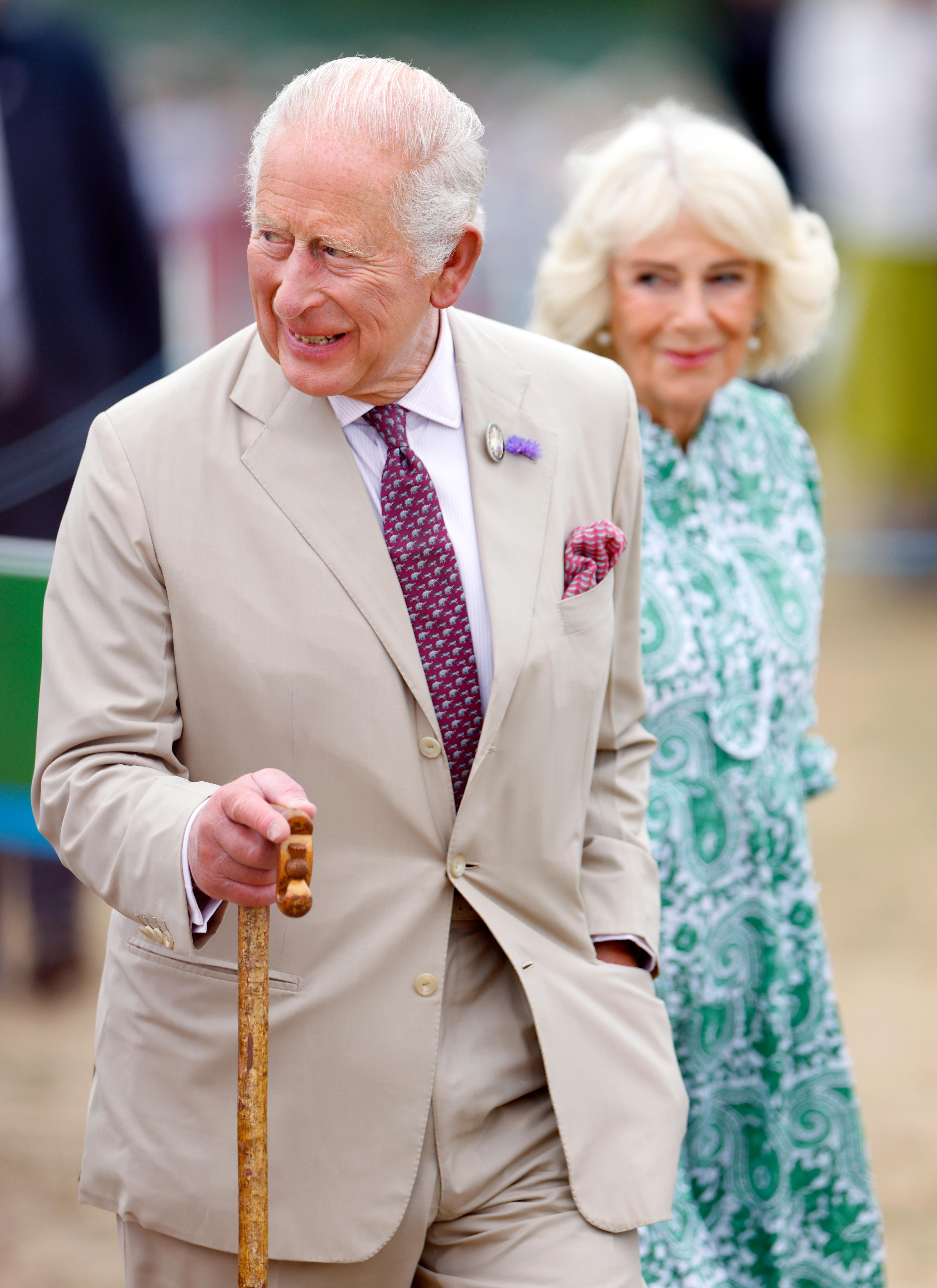 King Charles wears a light tan suit and carries a wooden walking stick while attending the RHS Sandringham Flower Show
