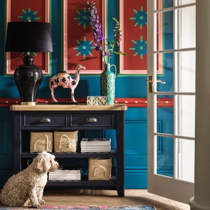 hallway wall decor ideas, blue hallway with handprinted paneling, navy painted console with lamp, vase, ornaments, wooden floor, rug, open door 