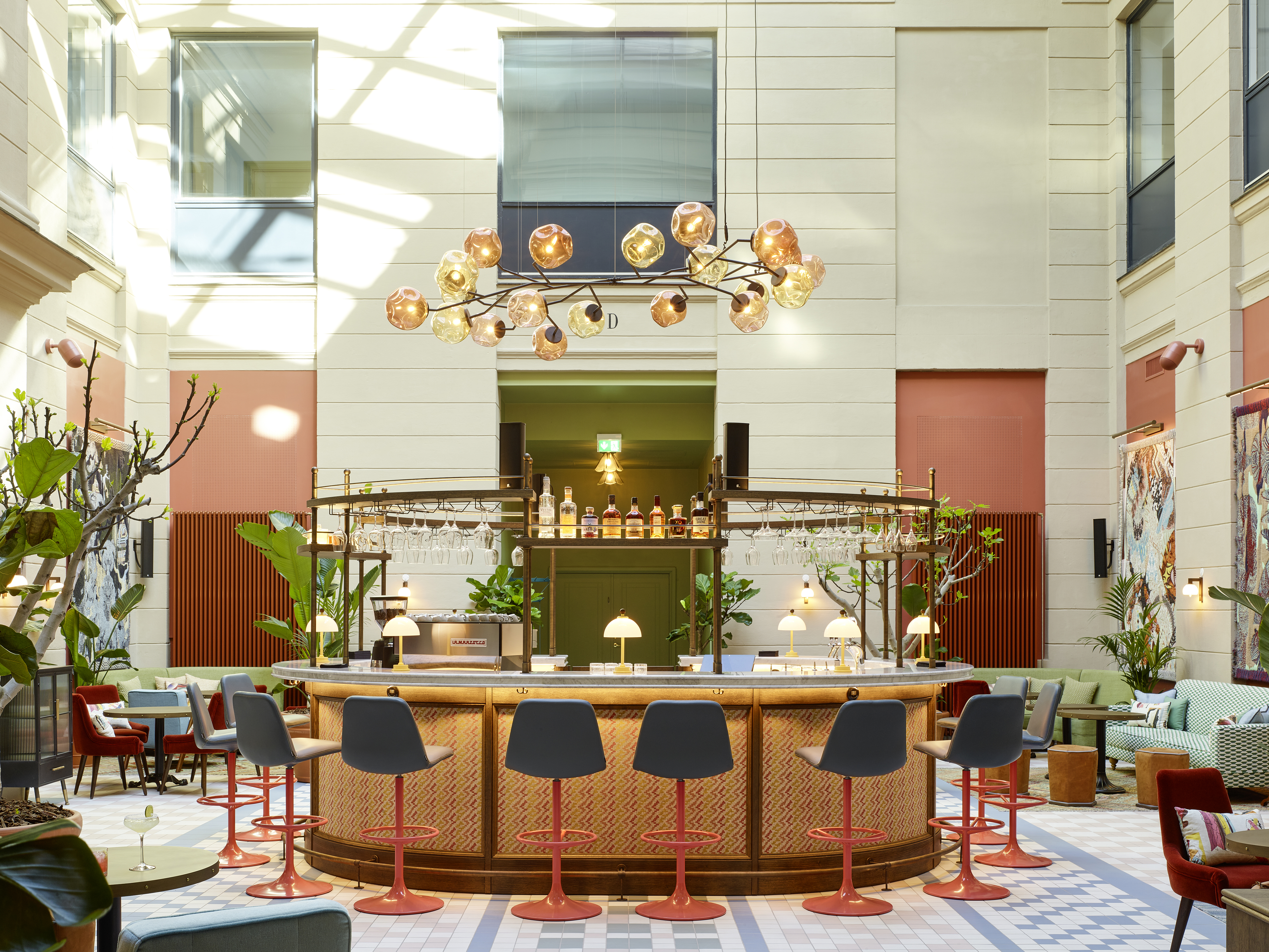 A colorful hotel inner courtyard boasts Space Age-inspired stools in blue textile and orange plastic, positioned around a circular bar decorated with mushroom-shaped table lamps, a multi-arm chandelier with spherical lights, a mosaiced floor, and a pastel palette.