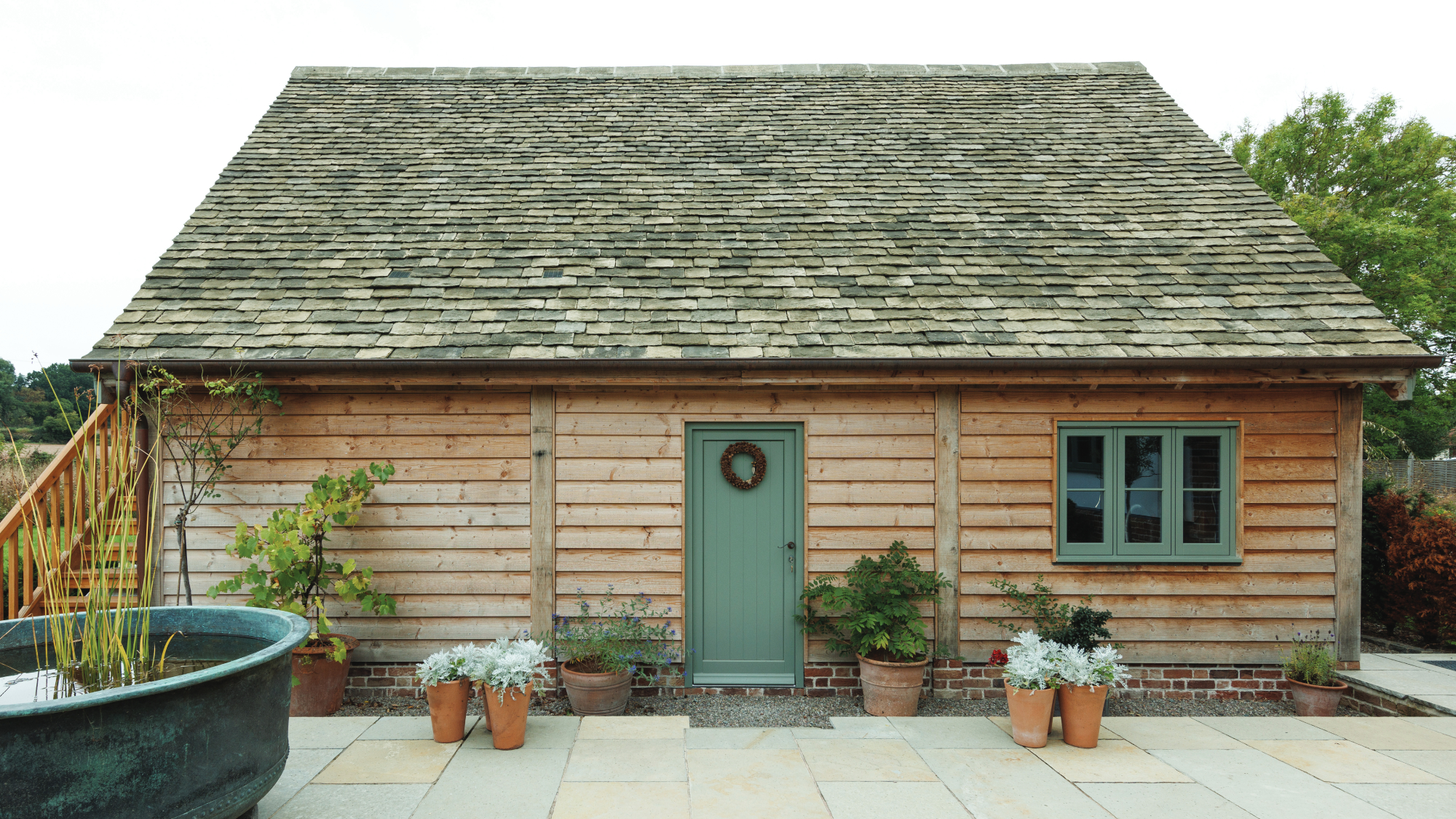single storey timber clad building with green door and green window leading onto tiled patio area
