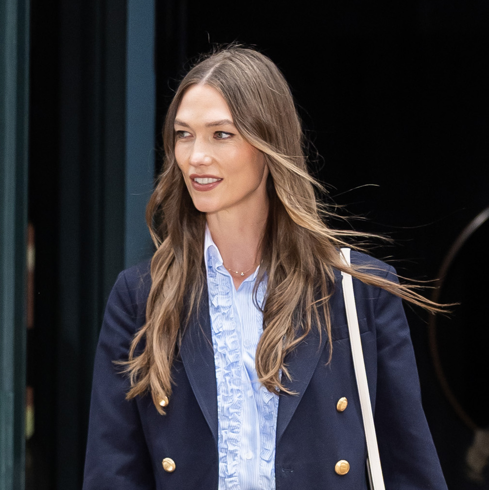 Karlie Kloss wearing a navy blazer over a ruffled blue button-down skirt with a frayed-hem denim midi skirt and brown suede ballet flats.