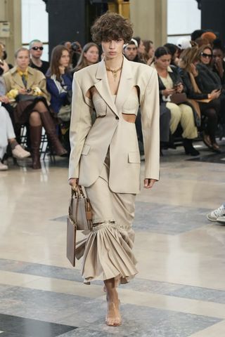 A female model wearing a light brown long sleeved dress with a handbag in her hand.