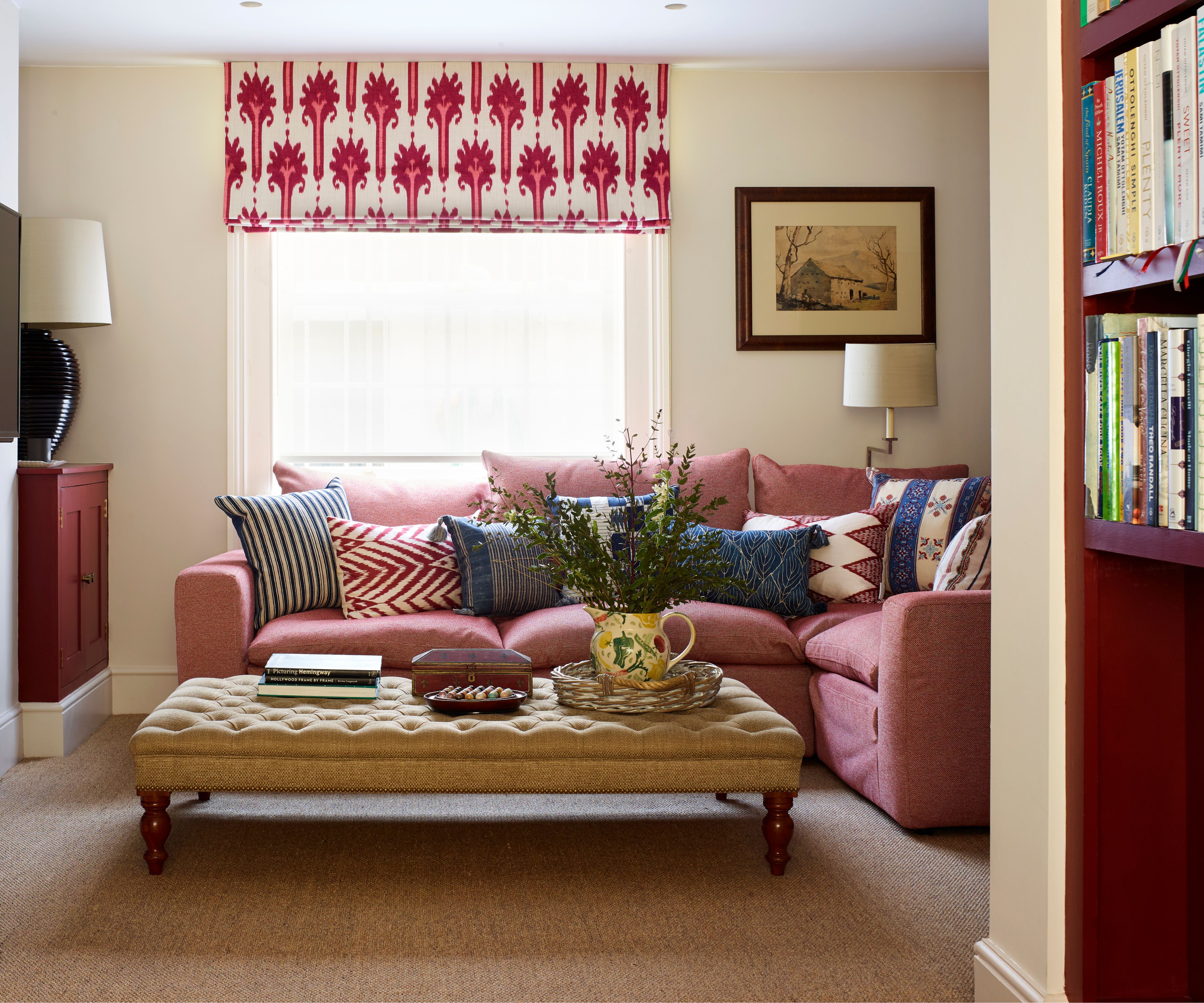neutral living room with a pink patterned window blind, pink corner couch with mix and match patterned pillows, a yellow ottoman, and red burgundy painted shelving and cabinetry