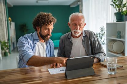 Adult son assisting elderly father with digital tablet and paperwork, in a comfortable home setting.