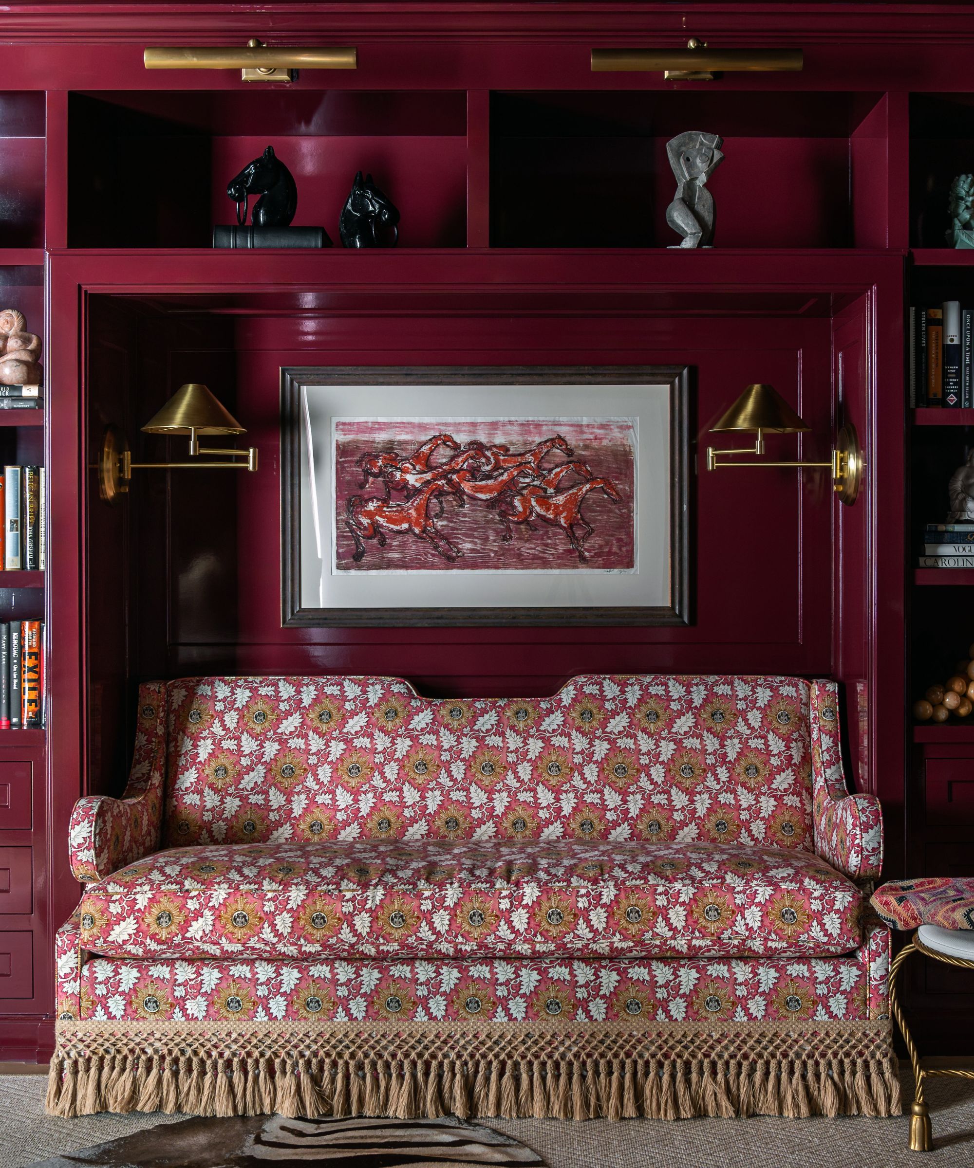 Study with pink lacquer built-in shelving, a nook with a patterned loveseat and a painting of horses above, two gold sconces, and books and statues in the alcoves.