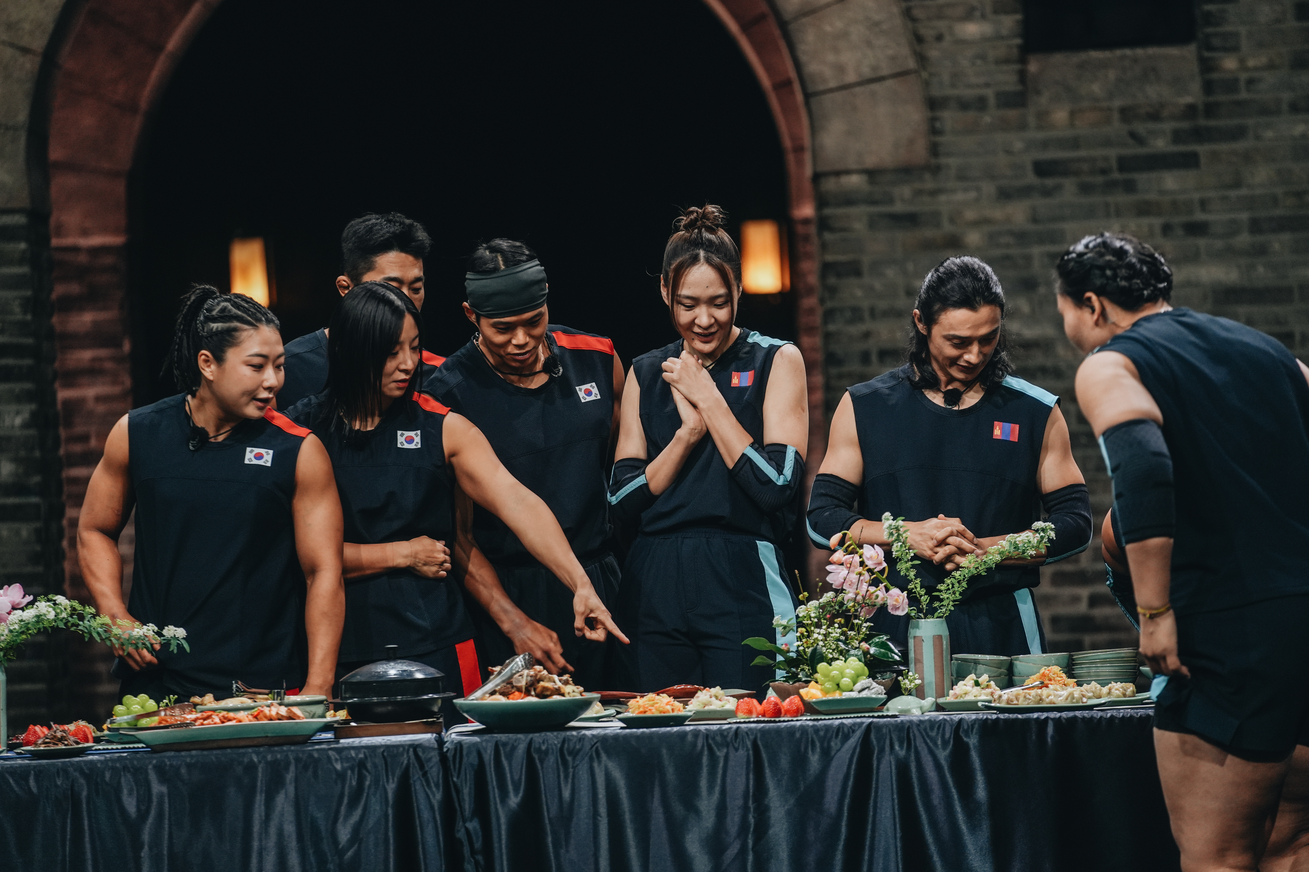 A group of athletes (l-r Choi Seung-yeon, Jang Eun-sil, Kim Dong-hyun, Amotti, Khandsuren Gantogtokh, and TK) look around a table of food, in Netflix&#039;s &#039;Physical: Asia.&#039;