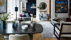 An eclectic open plan living and dining room, with a large black dining table in the foreground, various mismatched sofas and chairs with colorful cushions, and a large grey rug to the right with a large coffee table. The walls are white and dark blue, with mirrors, artwork and shelves hung up, and to the right is a large window that's filling the room with light.