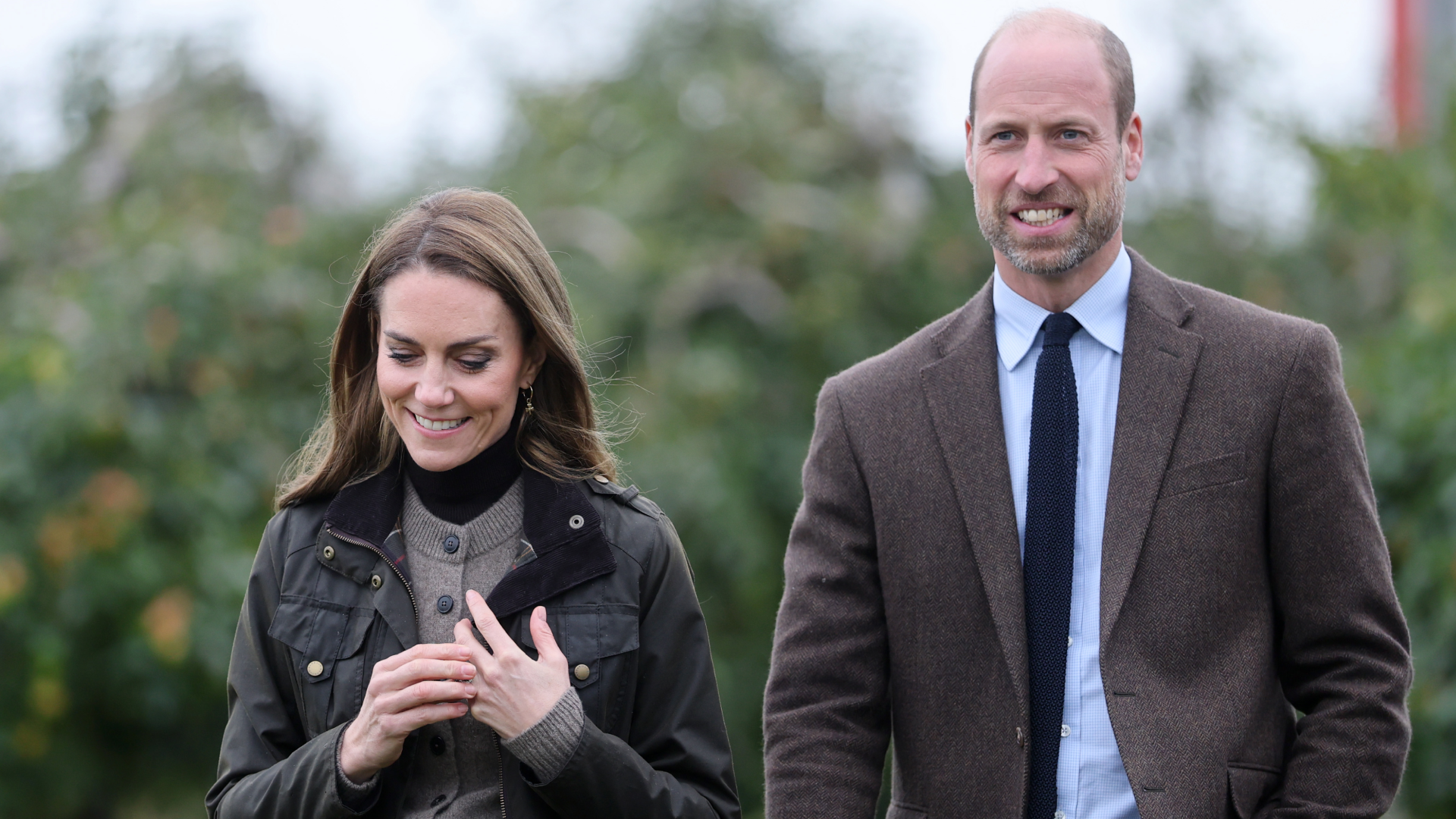 Prince William and Kate Middleton wearing brown and green jackets walking through a field