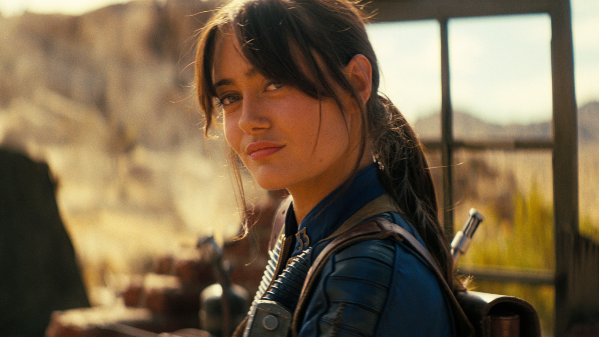 ella purnell in season 2 of fallout wearing the blue suit in a desert and smiling