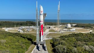 a red and white rocket rolls toward a launch pad, with trees and the ocean in the distance.