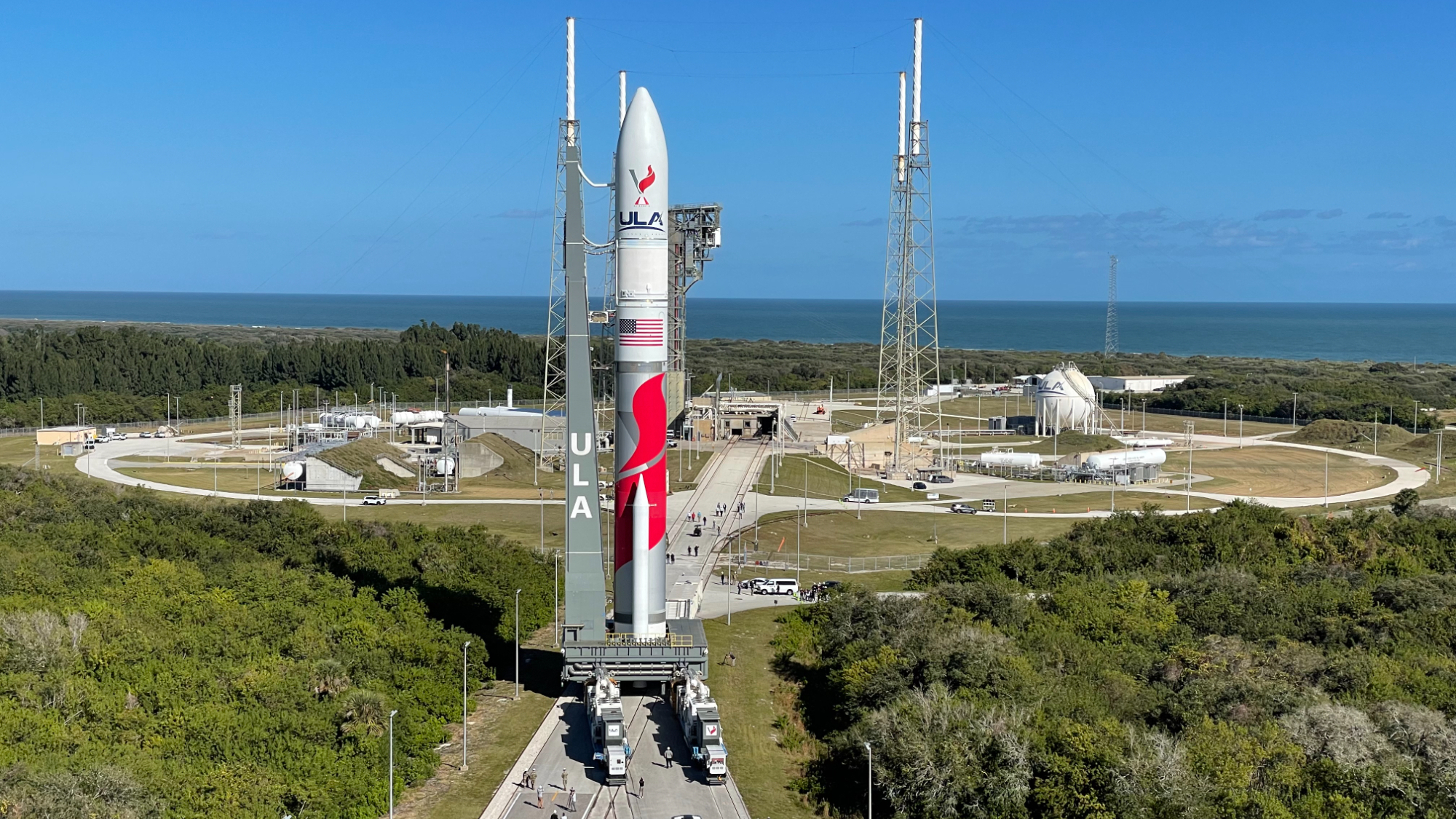 Red and white rocket standing on launch pad with ocean in background.