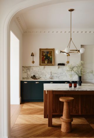 A west london kitchen with a dark wood island and a marble top, dark green cabinetry and a marble backsplash. Above the island is a gold light fixture