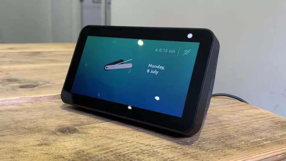 A black Amazon Echo Show 5 smart speaker on a wooden surface. The screen is showing the time and date.